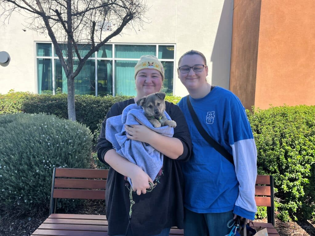 Two women smiling while holding a puppy wrapped in a blue blanket. They are standing outside near a bush and a wooden bench under a clear sky. The background includes a building with large windows. The image relates to Pet Adoptions.