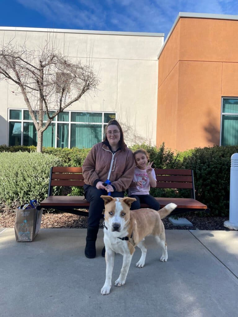 A woman and a young girl sit on a bench in front of a building, with a medium-sized dog standing in front of them. The scene is outdoors, featuring shrubs and a clear blue sky. This image highlights Pet Adoptions.