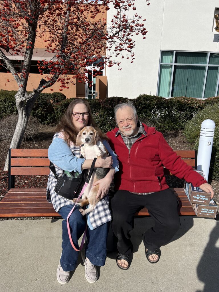 A woman in glasses and a plaid coat sits on a bench with a man in a red jacket. They are holding a small dog together. They are outside, with a few trees showing red leaves in the background. The image highlights the theme of Pet Adoptions.
