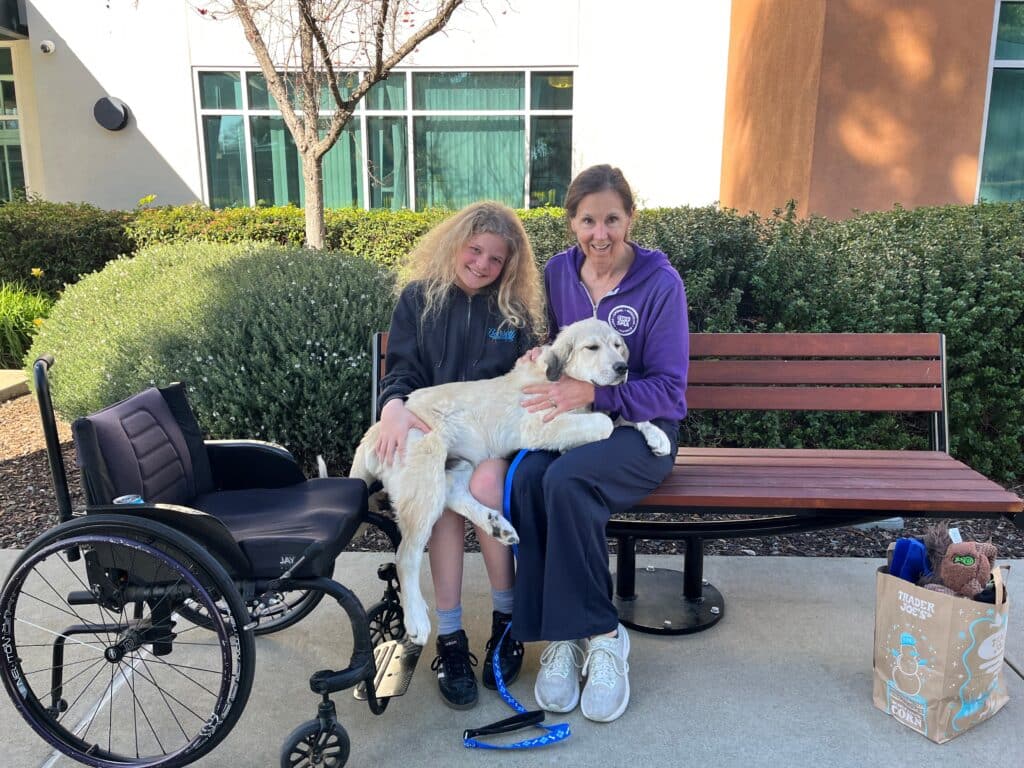A woman in a purple sweatshirt sits on a bench holding a small dog, while a girl with long hair sits beside her. A wheelchair is nearby, and there is a bag and leash on the ground. The setting includes green bushes and windows in the background, highlighting a moment of joy related to pet adoptions.