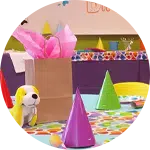 A colorful birthday party setup featuring several party hats in vibrant colors, a decorated table with a festive tablecloth, and a gift bag with pink tissue paper. A stuffed dog toy sits on the table, adding a playful touch.