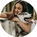 A young woman with long hair is hugging a gray and white dog with a leash attached. The scene is set outdoors with greenery in the background, suggesting a warm and friendly atmosphere.