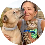 A woman smiles as a dog licks her face while she holds a cat wearing a blue and green patterned bandana. The background is a blurred outdoor setting, suggesting a pet-friendly environment.