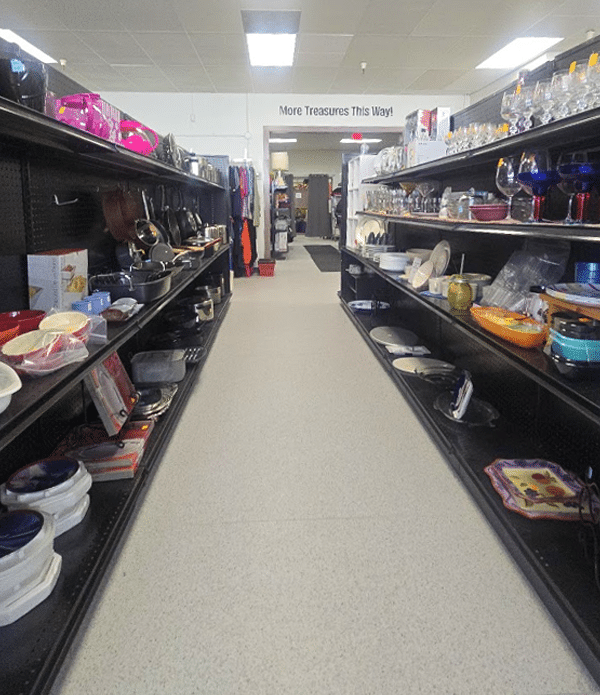 An interior view of the Joybound thrift store, showcasing aisles filled with various items like dishware, kitchenware, and decorative pieces. The space is organized with shelves on either side and a sign indicating 'More Treasures This Way' at the far end.