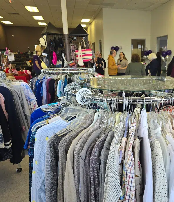 Inside Joybound thrift store, a display of clothes hangs on racks, with a focus on neatly arranged shirts. In the background, customers engage in conversation, and colorful balloons are visible, enhancing the lively atmosphere.