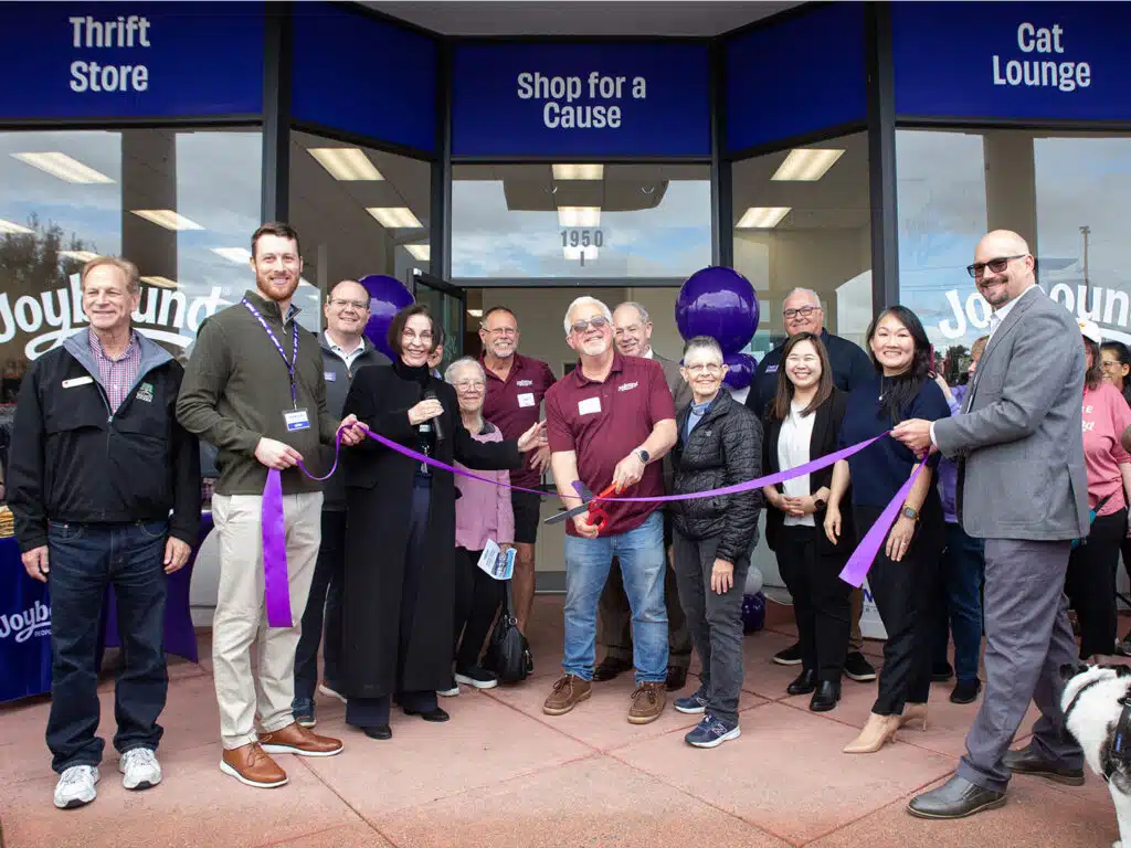A group of individuals, including men and women, stand outside a thrift store with a purple ribbon. They are smiling and holding the ribbon while preparing to cut it. The store's sign reads "Thrift Store," "Shop for a Cause," and "Cat Lounge."