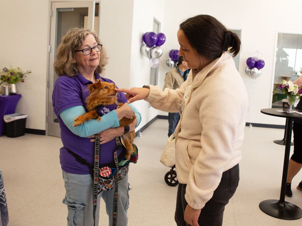 A woman with curly hair holding a small dog in her arms while another woman reaches out to pet the dog. They are inside a brightly lit space with purple balloons and floral arrangements in the background.