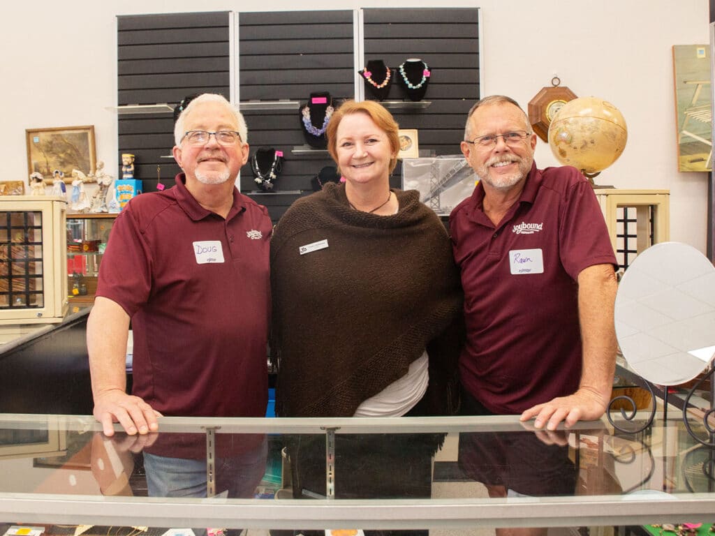 Three people standing behind a display counter in a thrift store. Two men on either side are wearing maroon shirts with name tags, and the woman in the center is wearing a dark shawl. The background includes shelves with various items.