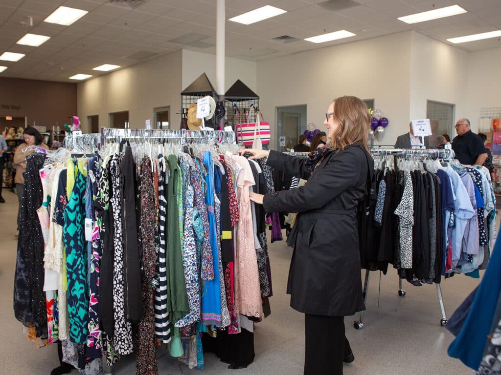 A woman in a black jacket browsing through racks of clothing at a thrift store. The store features a variety of dresses and tops displayed on metal hangers, with shoppers visible in the background. Bright lighting illuminates the spacious, organized space.