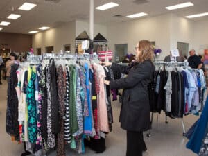 A woman in a black jacket browsing through racks of clothing at a thrift store. The store features a variety of dresses and tops displayed on metal hangers, with shoppers visible in the background. Bright lighting illuminates the spacious, organized space.