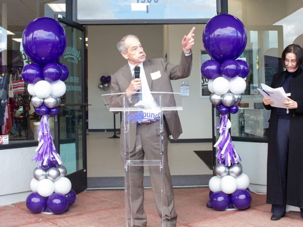 A man in a suit stands at a podium outdoors, speaking with his hand raised. To his left, a woman holds a document and stands next to decorated columns of purple, white, and silver balloons, marking the entrance of a thrift store during its re-opening event.