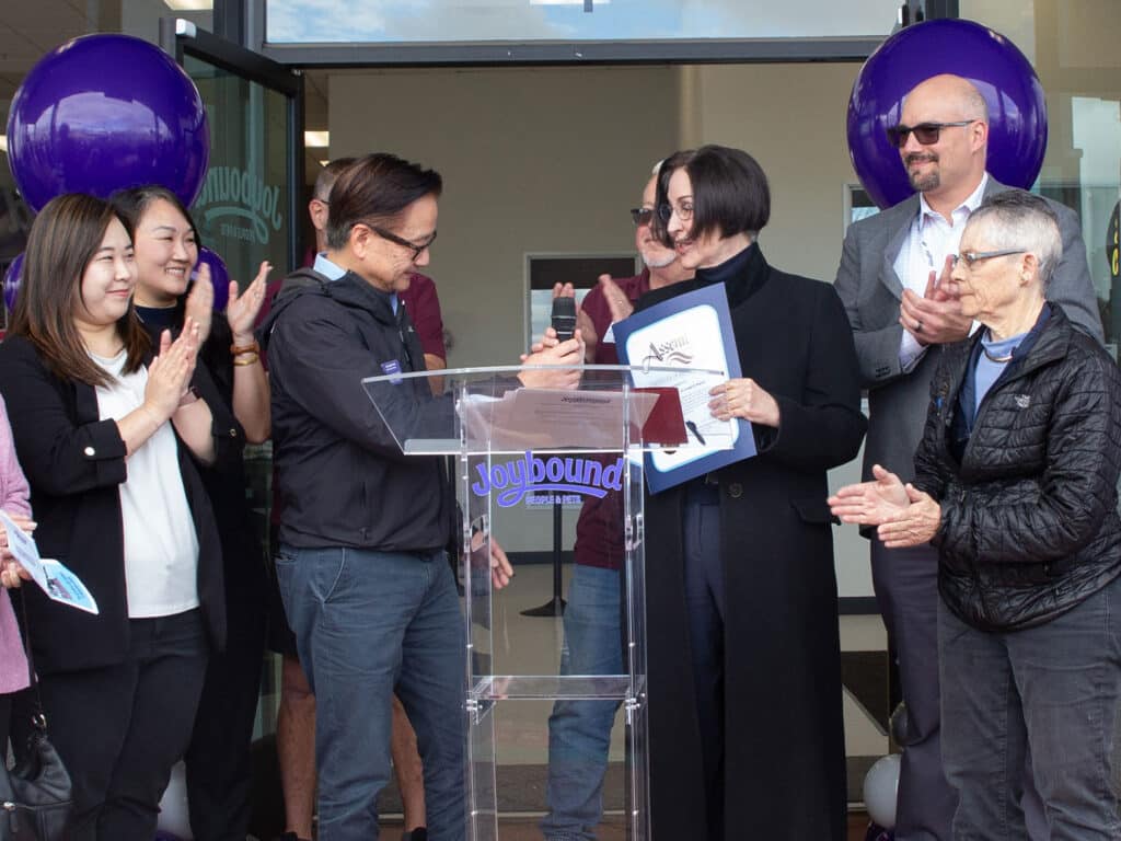 A group of people at a thrift store re-opening event, with two individuals at the front exchanging a certificate. Attendees are smiling and clapping, with purple balloons in the background. The atmosphere appears celebratory.