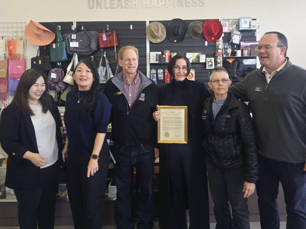 A group of six people stands together in a thrift store. They are smiling and posing with an official certificate. Behind them is a display of various hats and bags. The store has a sign that reads "UNLEASH HAPPINESS".