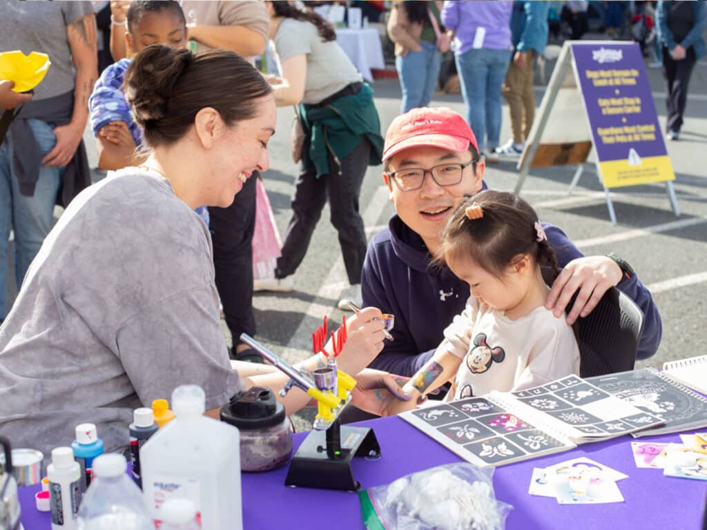 A woman in a gray top smiles while painting a child's face at a colorful outdoor event. A man in a red cap sits nearby, gently holding the child who appears to be around five years old, showing interest in the face painting.