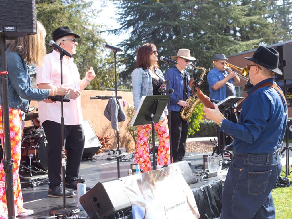 A live music performance featuring a band on stage. The vocalist stands at the microphone with two backup singers, while a guitarist and saxophonist play. The musicians wear colorful outfits and hats, and there's a backdrop of trees.