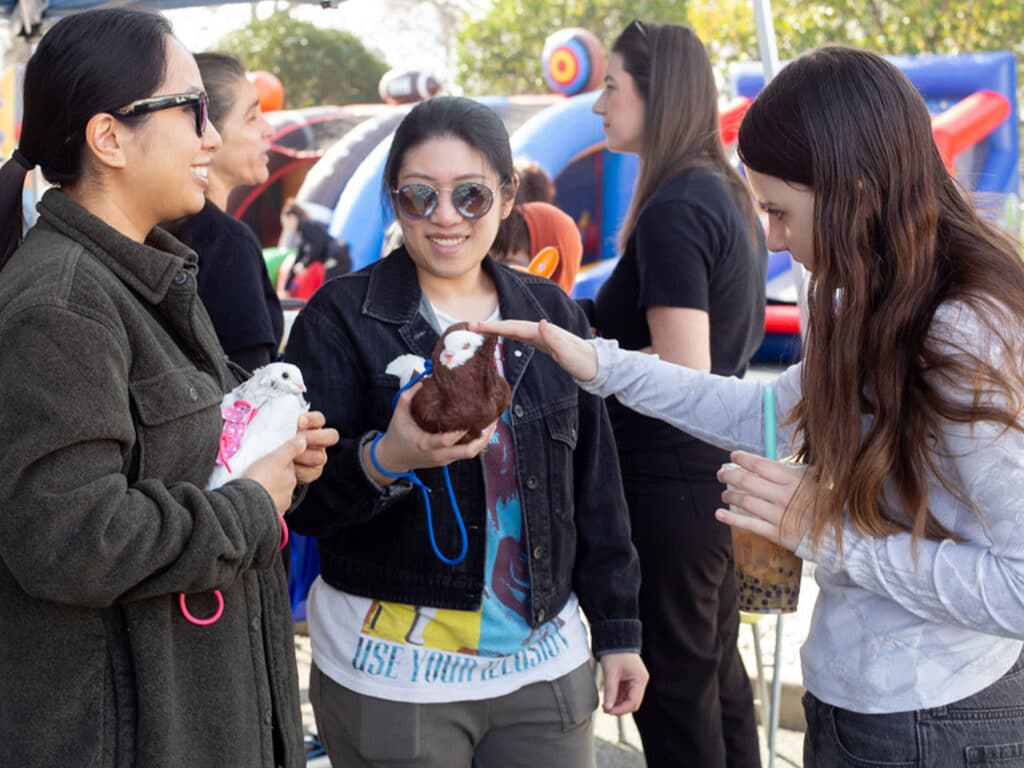Three women interact in an outdoor setting. One woman holds a small white bird, another has a brown plush animal, and the third gently touches the plush. A colorful inflatable structure is visible in the background.