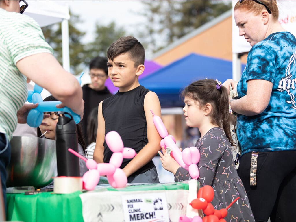 Two children, a boy wearing a sleeveless shirt and a girl with ponytails, stand at a booth receiving balloon animals. An adult is seen in a green striped shirt handing out the balloons. Several tents are visible in the background.