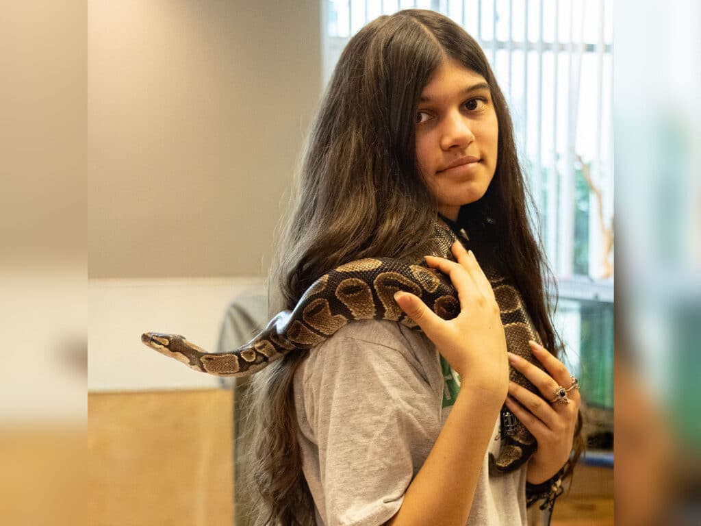 A young woman with long dark hair stands in a room, holding a snake draped over her shoulder. She wears a light-colored T-shirt and appears calm as she interacts with the snake, with natural light coming from a window in the background.