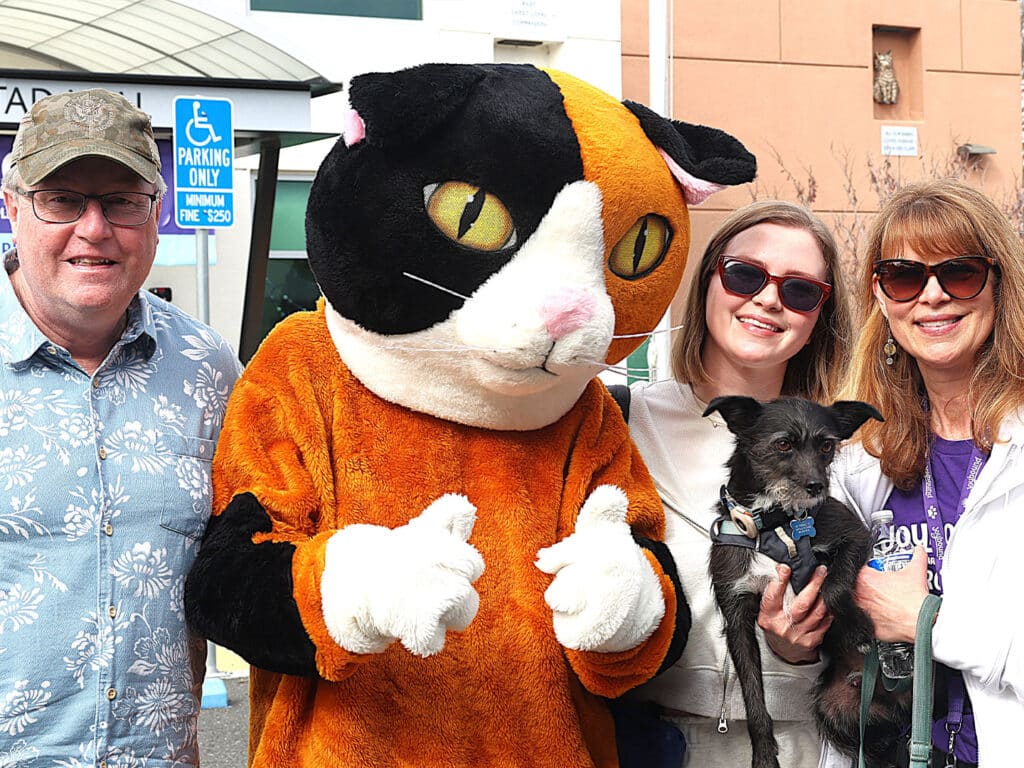 A group photo featuring a costumed cat character, two women, a man, and a small dog. The man, wearing glasses and a hat, stands on the left. The cat character is in the center, while the two women are on the right, one holding the dog.