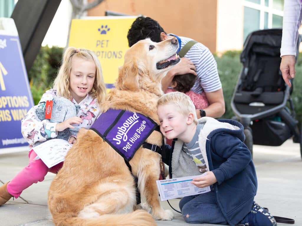 A golden retriever with a vest, labeled "Pet Hug Place," is surrounded by two children, a girl and a boy, who are smiling and interacting with the dog. A person in the background is helping another child. Several adoption signs are visible.