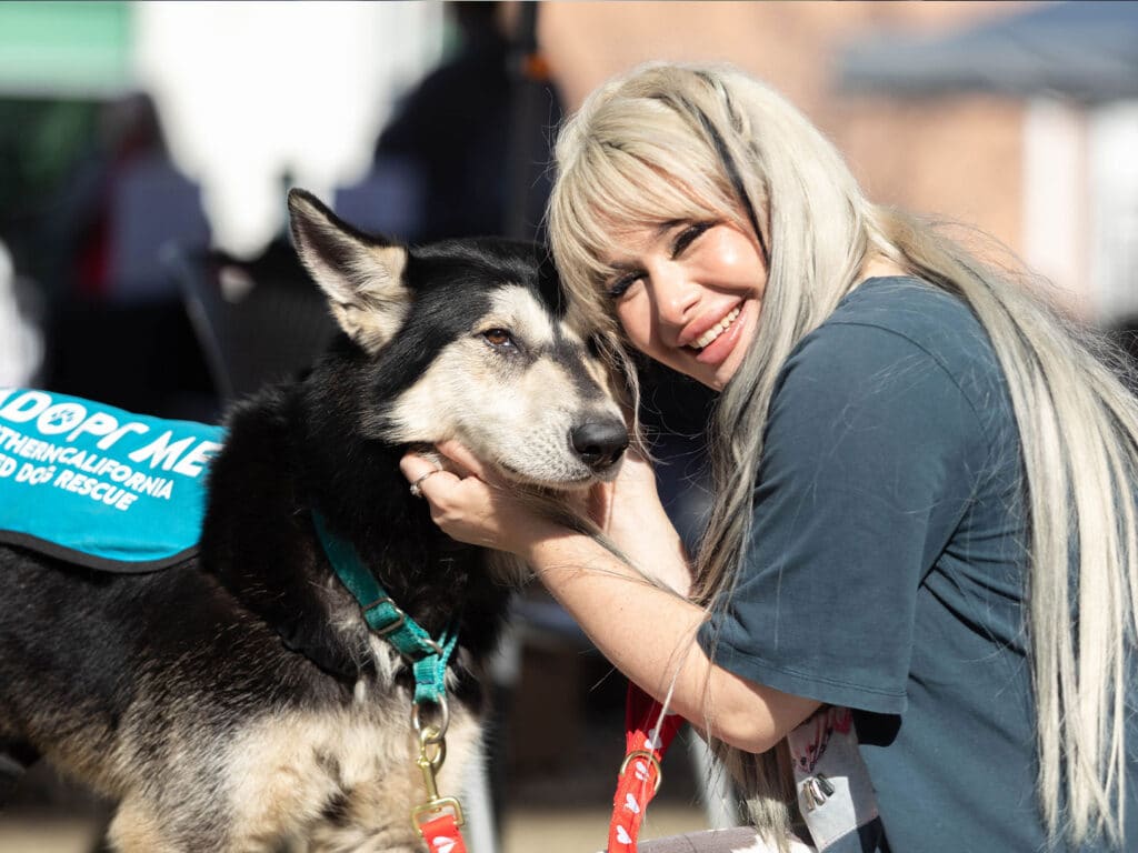 A woman with long blonde hair smiles while gently holding the face of a large black and tan dog. The dog wears a blue harness with the words "Adopt Me" on it. The background shows a bright outdoor setting with blurred figures.