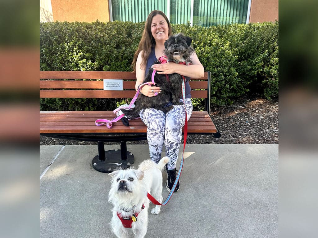 A woman sitting on a bench smiles while holding a small black dog in her lap. A white dog with a red harness stands on the ground beside her. The background shows green shrubbery and features a sign on the bench.