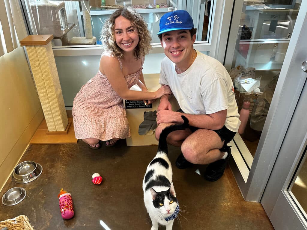 A woman in a light dress and a man in a white T-shirt are kneeling beside a cat in an animal shelter. The cat, with black and white fur, is standing in front of them on a concrete floor, while toys are visible nearby.