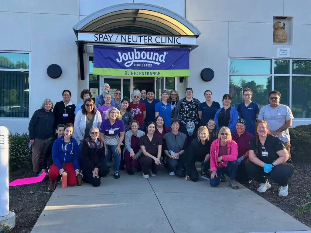 A large group of approximately 30 individuals poses together in front of the Spay/Neuter Clinic, smiling and dressed in various clinic uniforms. A banner with "Joybound" hangs above the entrance, indicating the event's cooperative spirit.
