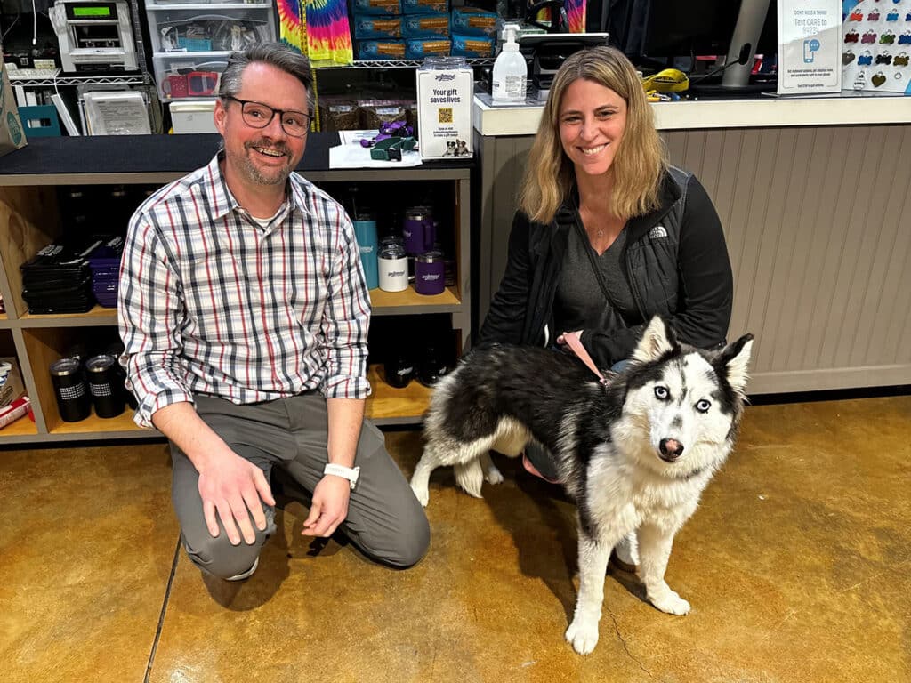 A man and a woman crouch on the floor beside a Siberian Husky in a store setting. The man, wearing glasses and a plaid shirt, smiles as he poses with the woman, who has long hair and wears a black jacket.