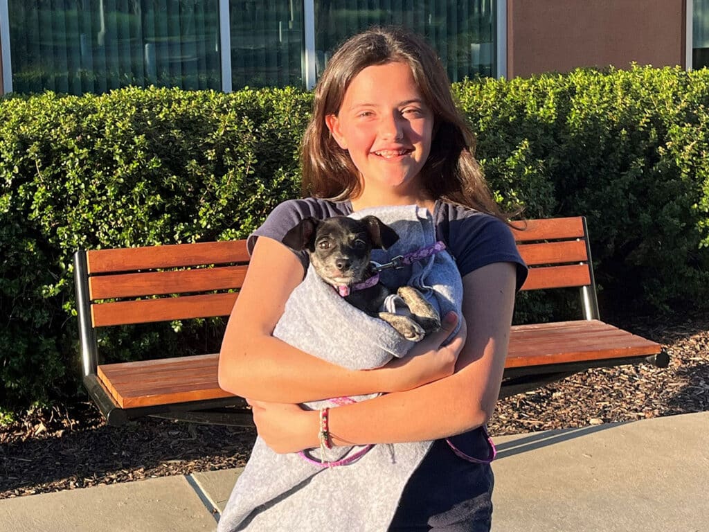 A girl with long brown hair smiles while holding a small dog wrapped in a gray blanket. They stand in front of a wooden bench and green shrubbery, with sunlight illuminating the scene.