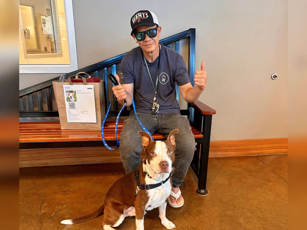 A man wearing sunglasses and a baseball cap sits on a bench, giving a thumbs-up. Next to him is a brown and white dog on a leash, looking at the camera. Behind them is a wooden partition and a bag on the floor.