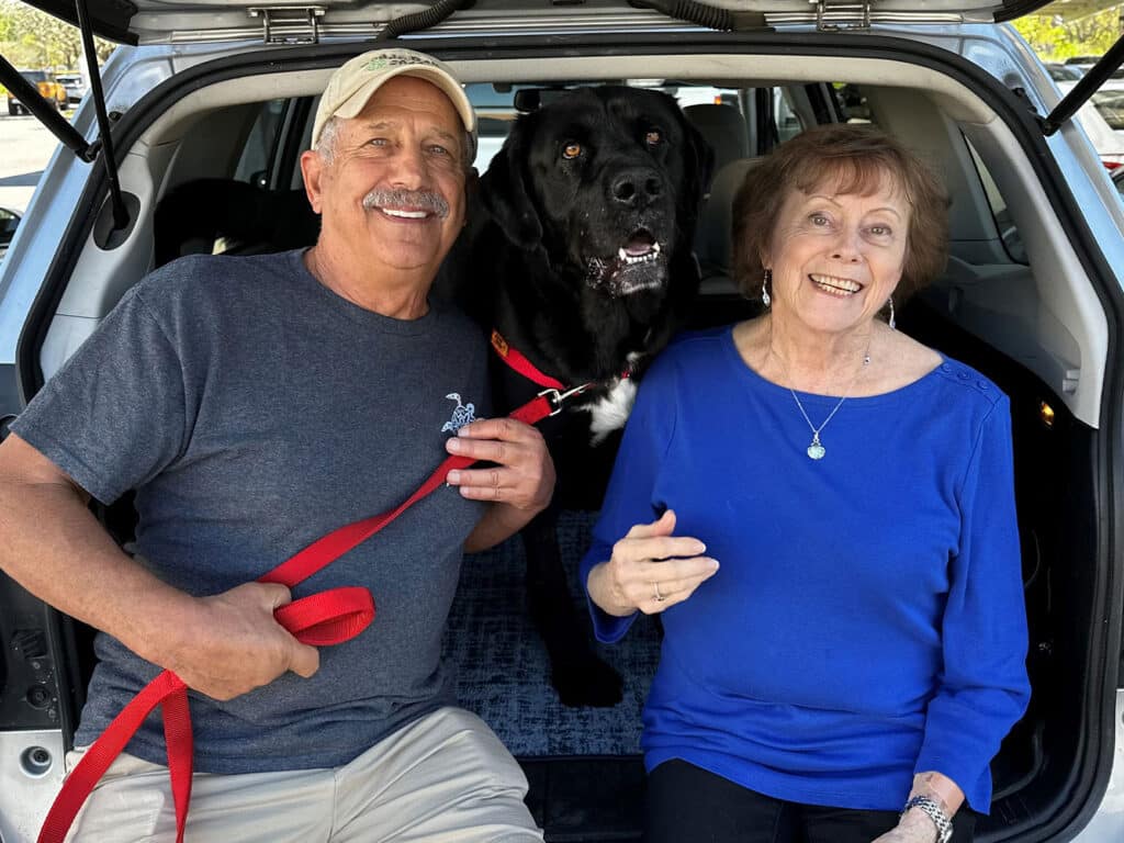 A man and a woman sitting in the back of an SUV with a large black dog. The man is wearing a gray T-shirt and a cap, holding the dog's leash. The woman is in a blue top, smiling at the camera. The scene is bright and cheerful.