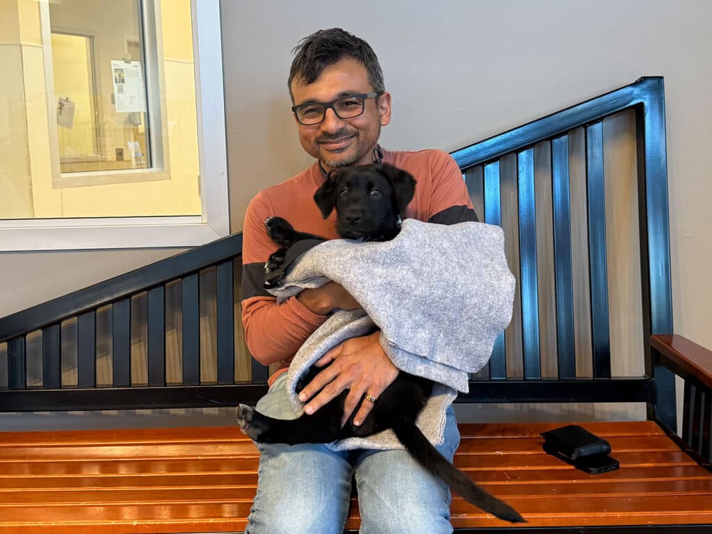 A man sitting on a wooden bench, holding a black puppy wrapped in a gray blanket. He is smiling and wearing glasses, dressed in a long-sleeve shirt and jeans. In the background, there is a window showing part of a room.