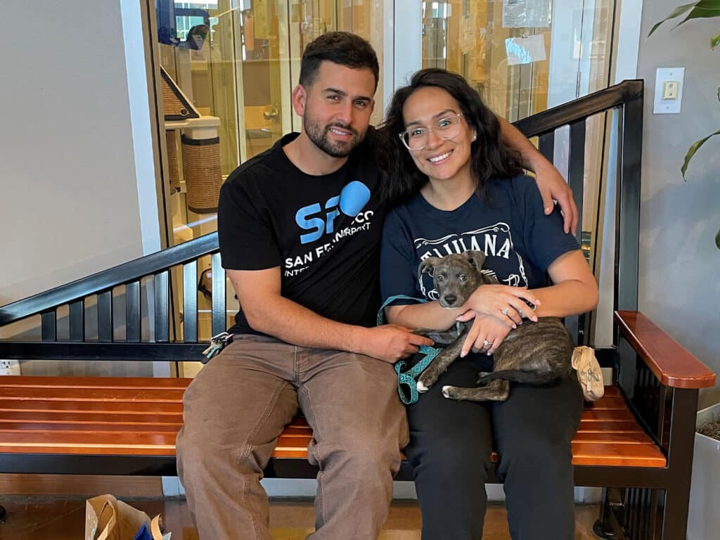 A man and a woman sitting on a bench in a pet adoption center, both smiling. The woman holds a small, brown and white dog in her lap, while the man gently rests his arm around her. They are in a bright, open space with plants in the background.