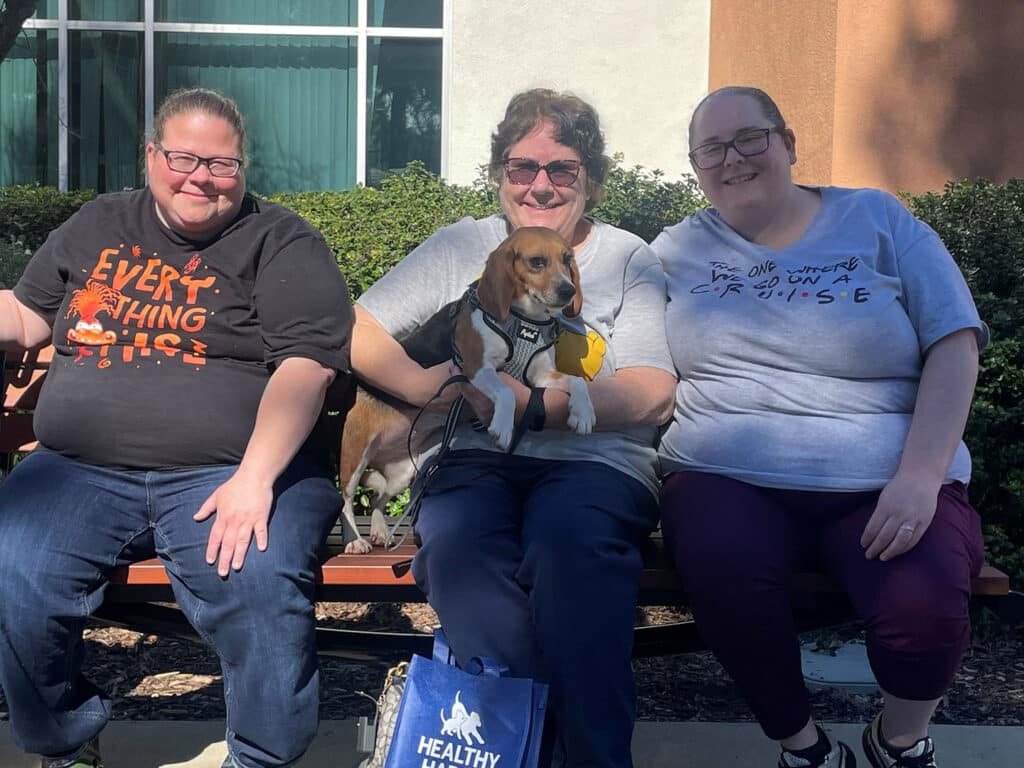 Three women sit on a bench outdoors, smiling. The woman in the middle holds a small dog. The leftmost woman wears a black shirt with "Everything This" text, and the rightmost woman is in a light blue shirt with text. A healthy-themed bag is on the ground.