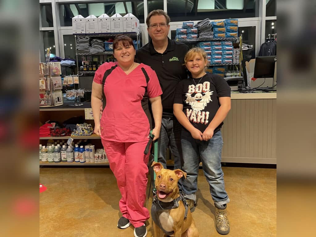 A woman in pink scrubs, a man in a black shirt, and a child in a black tee pose together with a brown dog in a pet adoption center. Shelves with pet supplies and products are visible in the background.