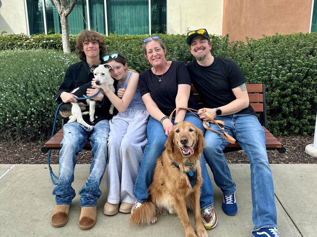 A family of four sits on a bench with two dogs. The mother holds a small dog, while the children are next to her. The father sits on the right with a golden retriever. They are outdoors, with green bushes in the background and a building in sight.