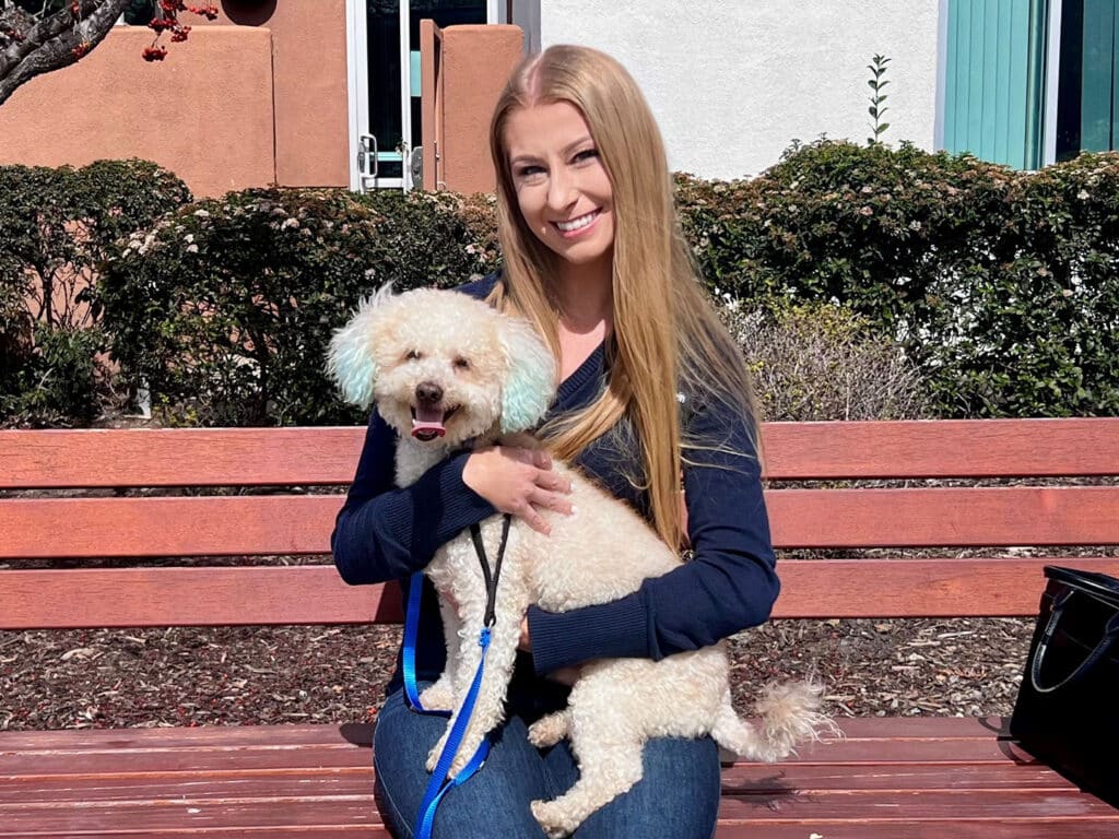 A smiling woman with long hair in a dark sweater sits on a wooden bench, holding a small cream-colored dog with light blue ears. Green shrubs and a building are visible in the background.