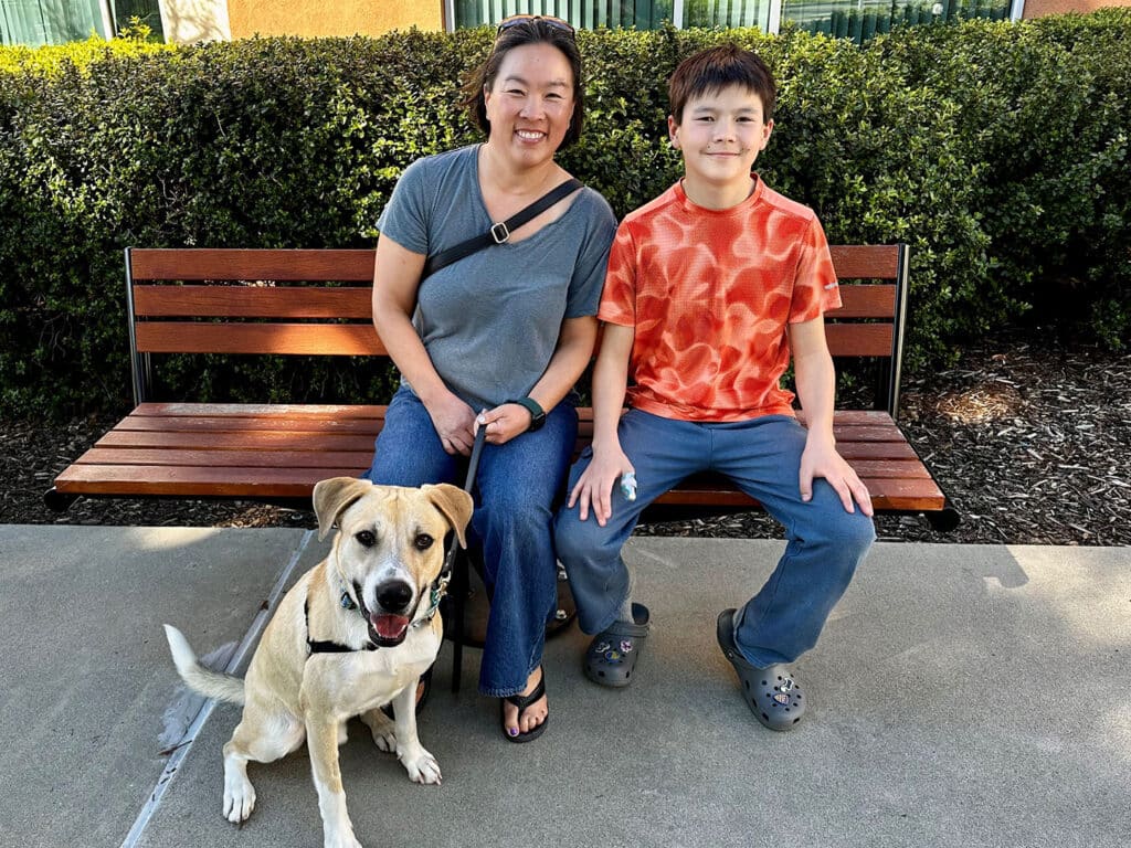 A woman and a boy sit on a wooden bench in a park. The woman has short hair and wears a gray shirt and denim jeans. The boy, wearing an orange and white patterned shirt and blue pants, sits beside her. A tan and white dog is on a leash next to them.