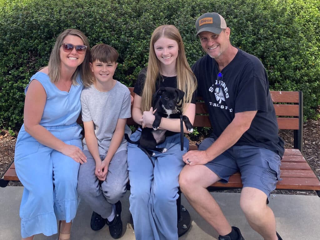 A family of four consisting of two adults and two children sits together on a bench in a park. The girl holds a small black dog, while the others smile at the camera, surrounded by greenery.