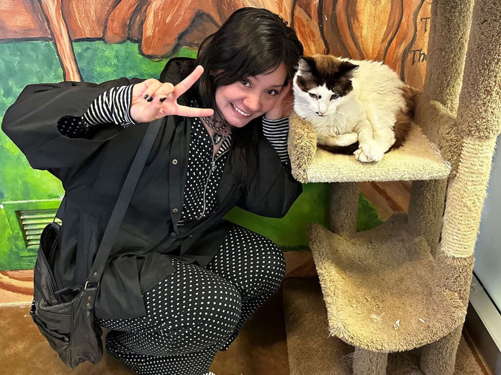 A woman with dark hair, dressed in a black and white polka dot outfit, poses peace sign next to a fluffy cat resting on a scratching post. The background features a vibrant mural, enhancing the pet adoption theme.