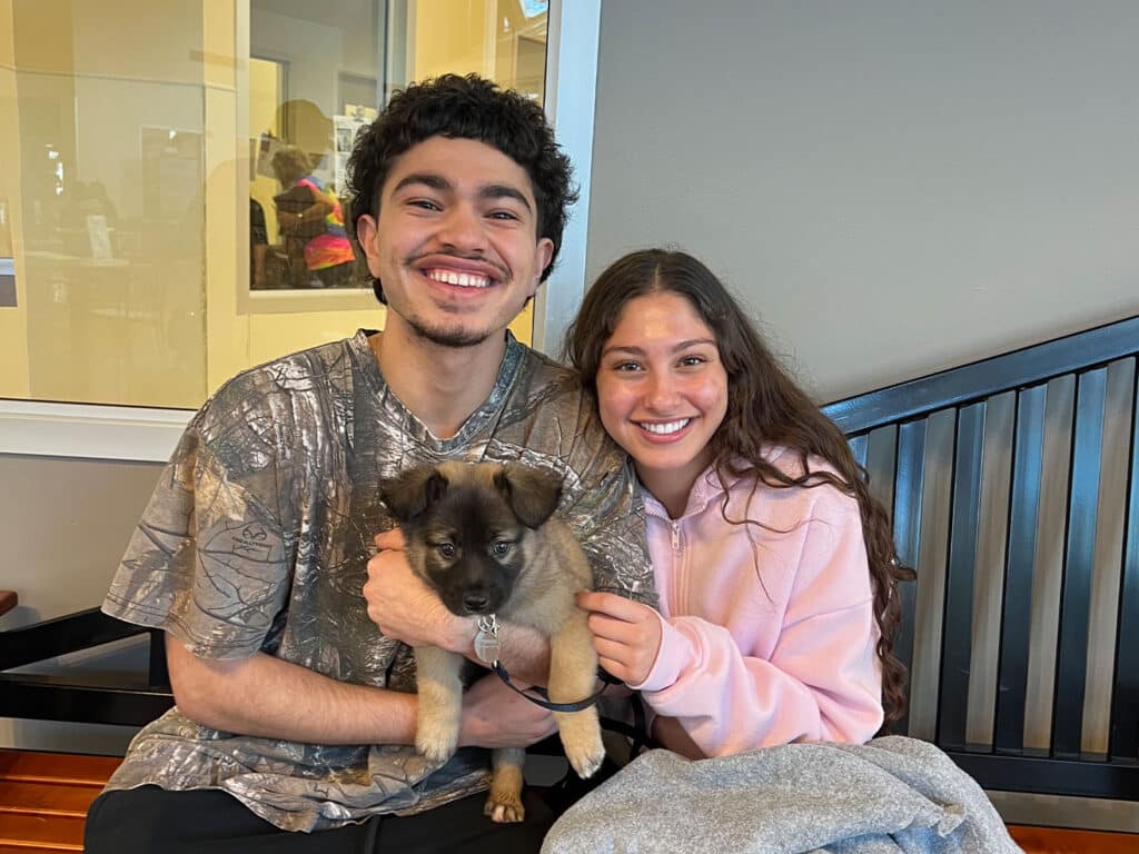 A young man and woman sitting together on a bench, holding a small puppy. The man has curly hair and is wearing a patterned shirt, while the woman is in a pink hoodie. They are smiling warmly at the camera, with a bright background visible.