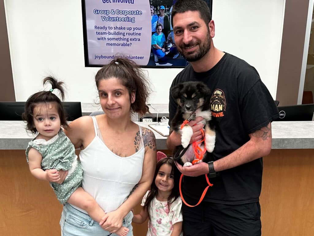 A man and a woman stand with three children and a small dog in a pet adoption center. The man holds the dog, while the woman carries a baby. Two girls stand beside them. A promotional poster behind them encourages volunteering.