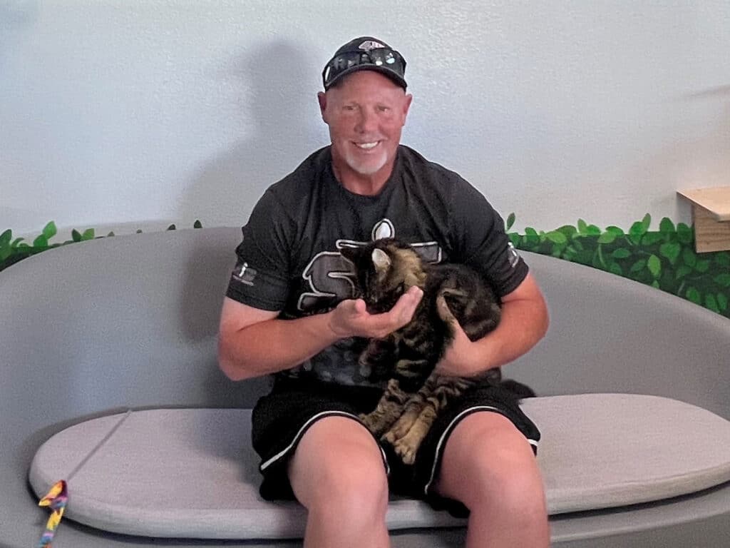 A man with a shaved head, wearing a black t-shirt and shorts, sits on a light gray couch holding a brown and black striped cat in his lap. The background features a green leafy design.
