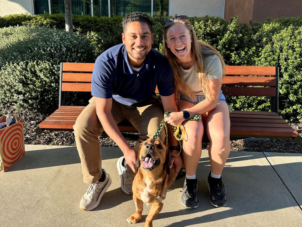 A man and a woman sit on a bench outside, smiling and posing with a brown dog on a leash. The man wears a navy polo shirt and khaki pants, while the woman is in a light shirt and shorts. Green bushes and a building are seen in the background.