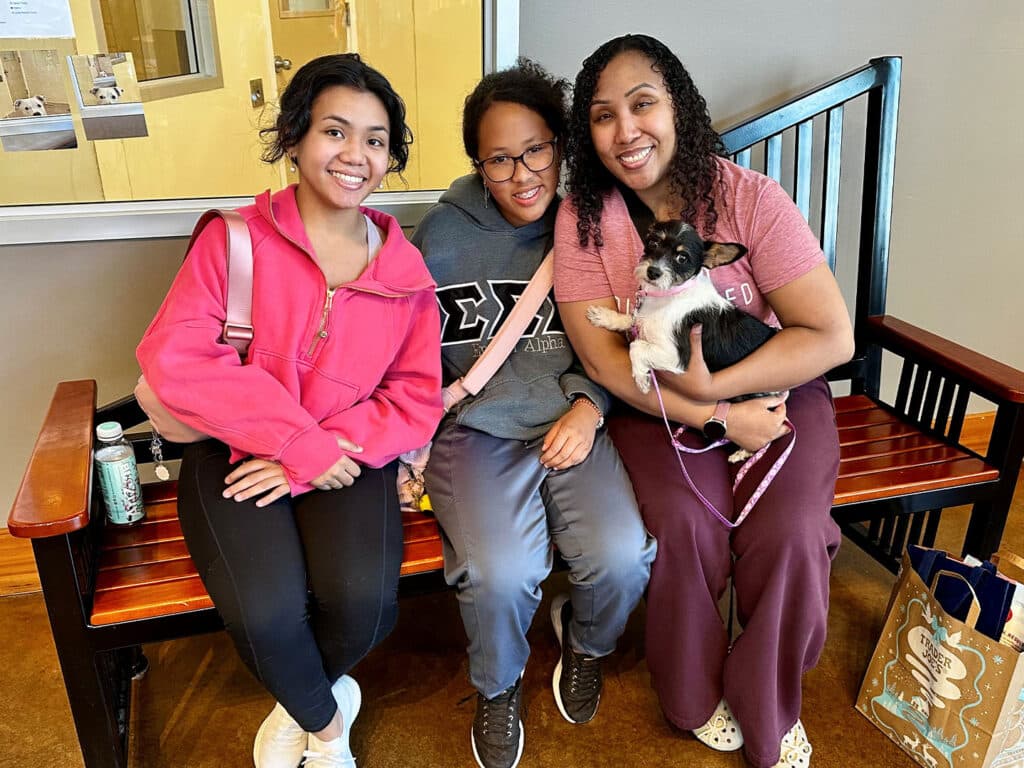 Three women sitting on a wooden bench indoors, smiling for the camera. One woman holds a small dog. The setting features warm colors and a wall with yellow and brown hues.