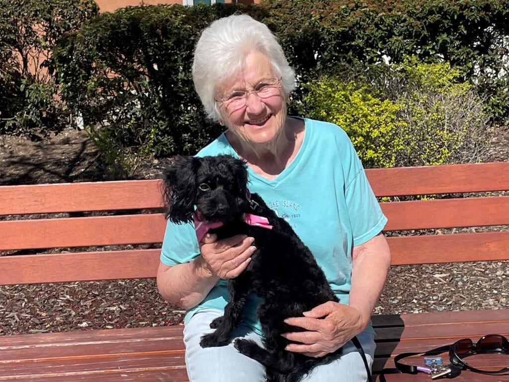 An elderly woman sitting on a wooden bench, smiling while holding a small black dog with a pink collar. The background features greenery and bushes, indicating a sunny outdoor setting.