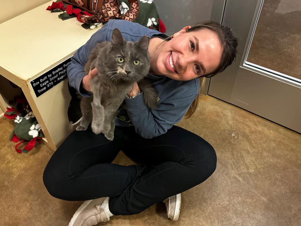 A woman sitting on the floor holding a gray cat. She smiles while looking at the camera, wearing a casual outfit. Behind her, there is a table with a blanket and a door leading to another room.