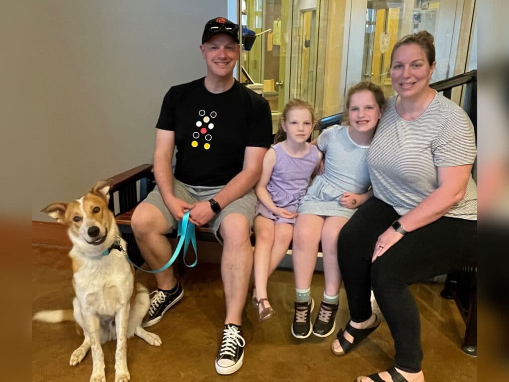 A family of four sits together on a bench with a dog. The man wears a black t-shirt and shorts, the woman in a striped shirt and black pants. Two girls, one in a purple dress and the other in a light blue shirt, smile beside them.