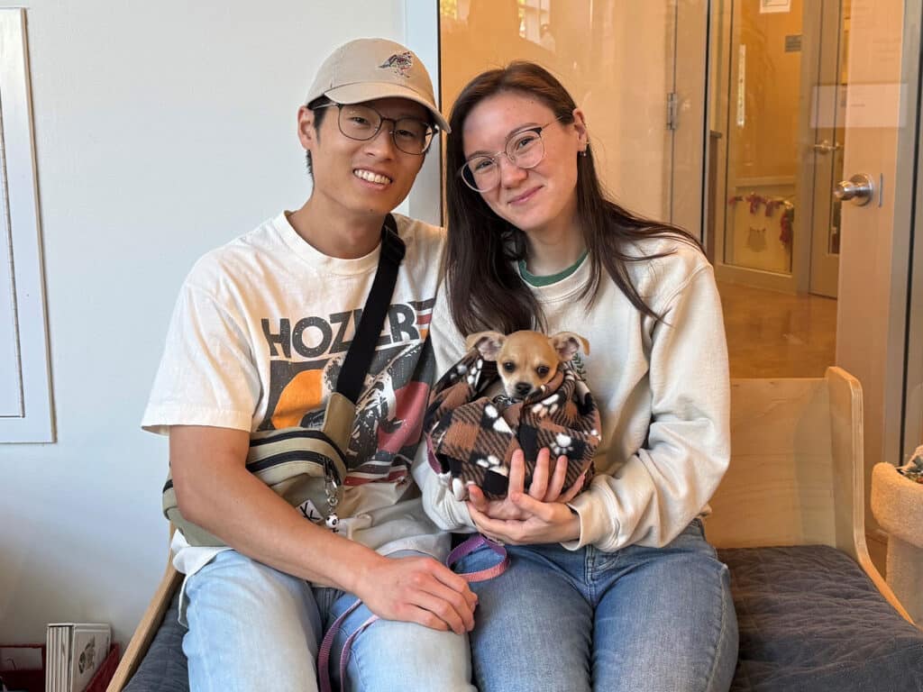 A young man and woman are seated on a bench in an adoption center. The man, wearing a cap and glasses, smiles alongside a woman in a sweatshirt and glasses, holding a small dog wrapped in a blanket.